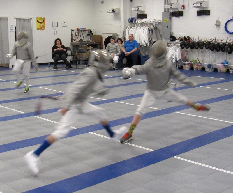Two fencers in full protective gear engage on an indoor fencing strip, with spectators watching in the background.